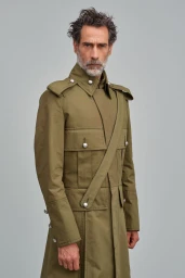 Middle-aged man with dark hair and grey beard wearing an olive green military-style coat against a plain grey background