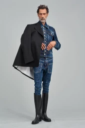Model wearing denim jacket and jeans with black boots holding a black coat against a grey background