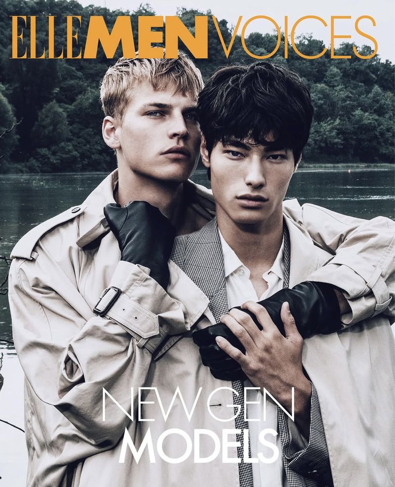 Two men pose in trench coats and black gloves against a blurred lake and forest backdrop for a magazine cover, captured in a medium shot.