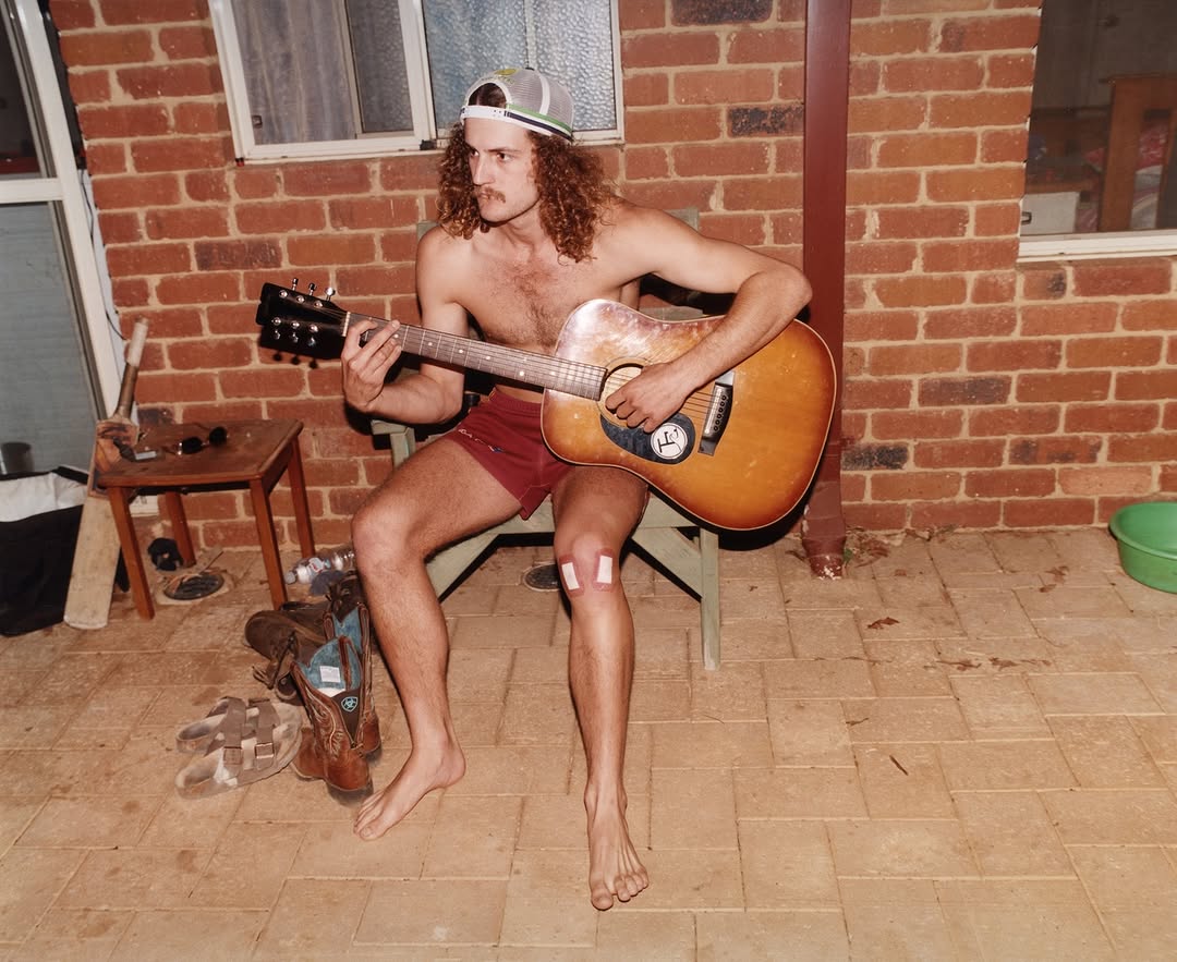 Shirtless man with long curly hair plays an acoustic guitar outdoors in front of a brick wall, wearing red shorts and a baseball cap.