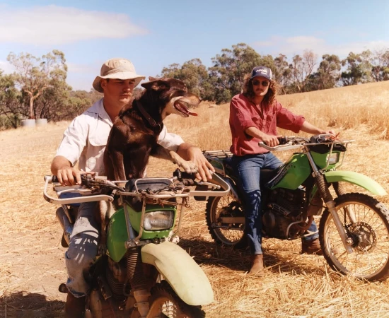 Two men are riding green dirt bikes in a grassy field on a sunny day, one man has a dog on his bike in front of him.