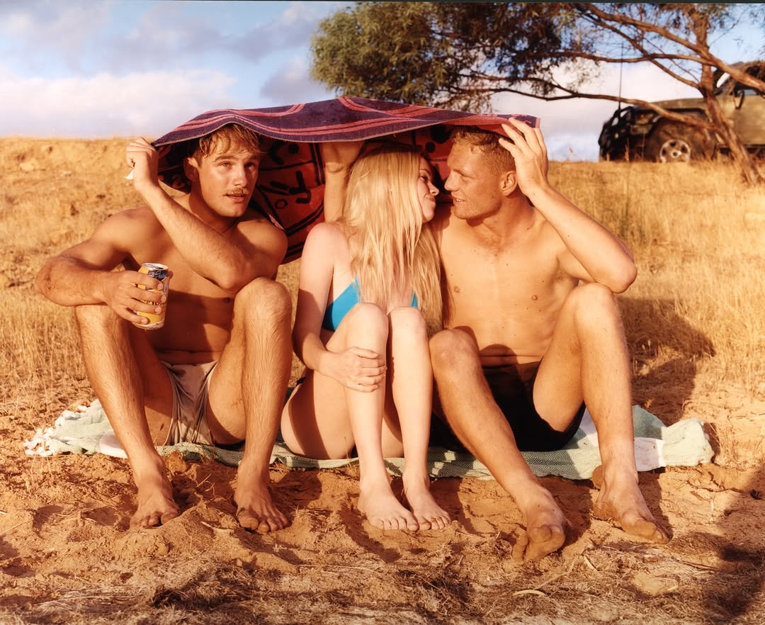 Three fair-skinned young adults sit together on a sandy hill under a striped towel, with a car and tree visible in the background.