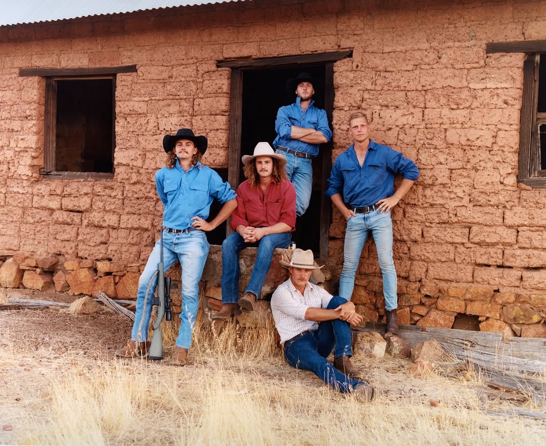 Five men in Western wear pose in front of a brick building in a rural area with dry grass and partial sunlight, evoking a vintage feel.