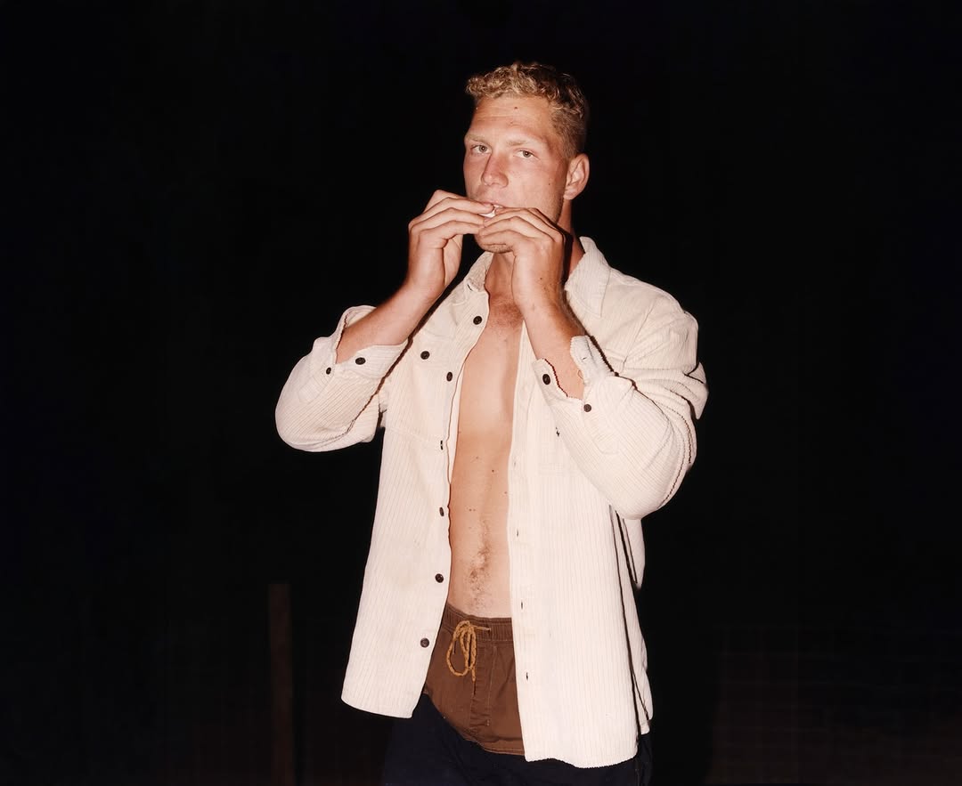 Young adult man in unbuttoned shirt holds hands near mouth as if playing a harmonica in a low-light outdoor setting.