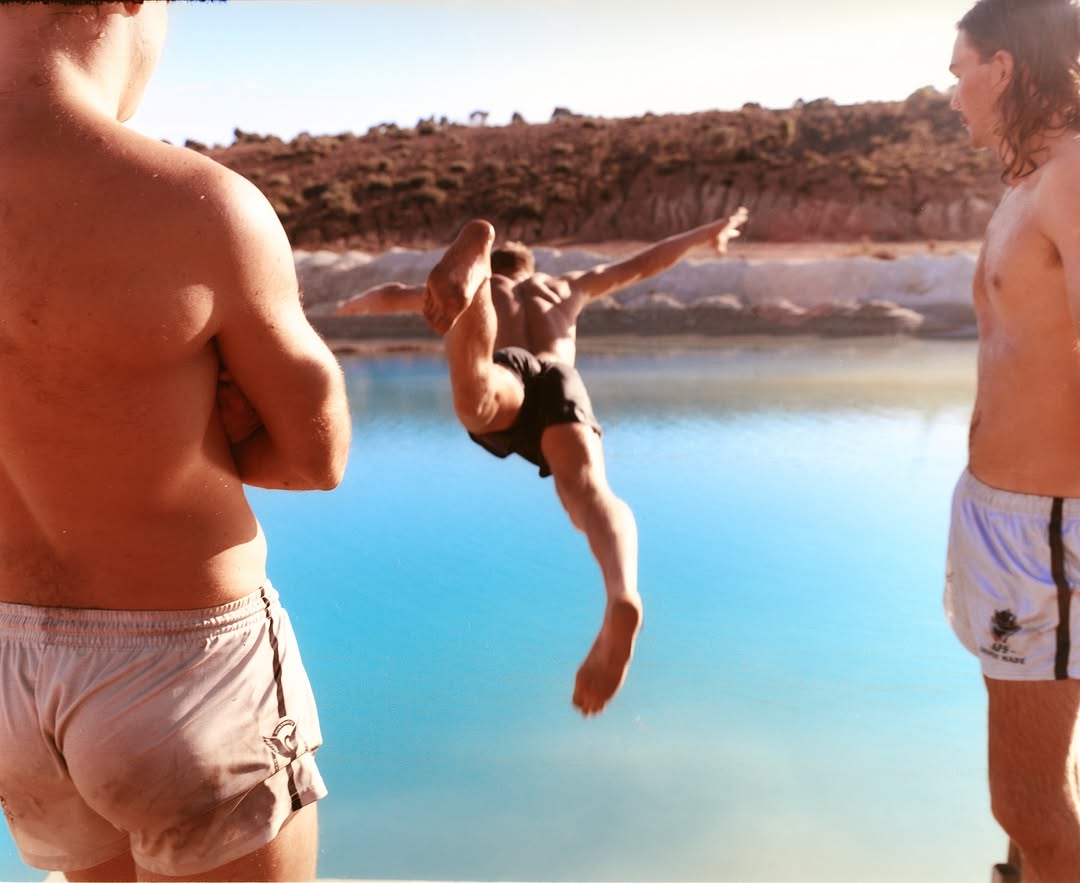 Man in mid-air dives into turquoise water, flanked by two shirtless men, against a backdrop of sparse, hilly terrain in bright daylight.