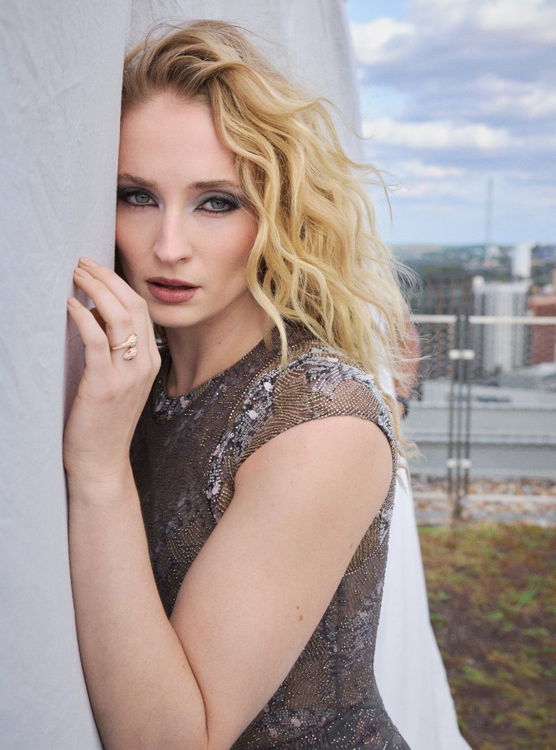 A portrait shot of a blonde woman with heavy eye makeup leaning against a textured white wall, with a cityscape visible in the background.