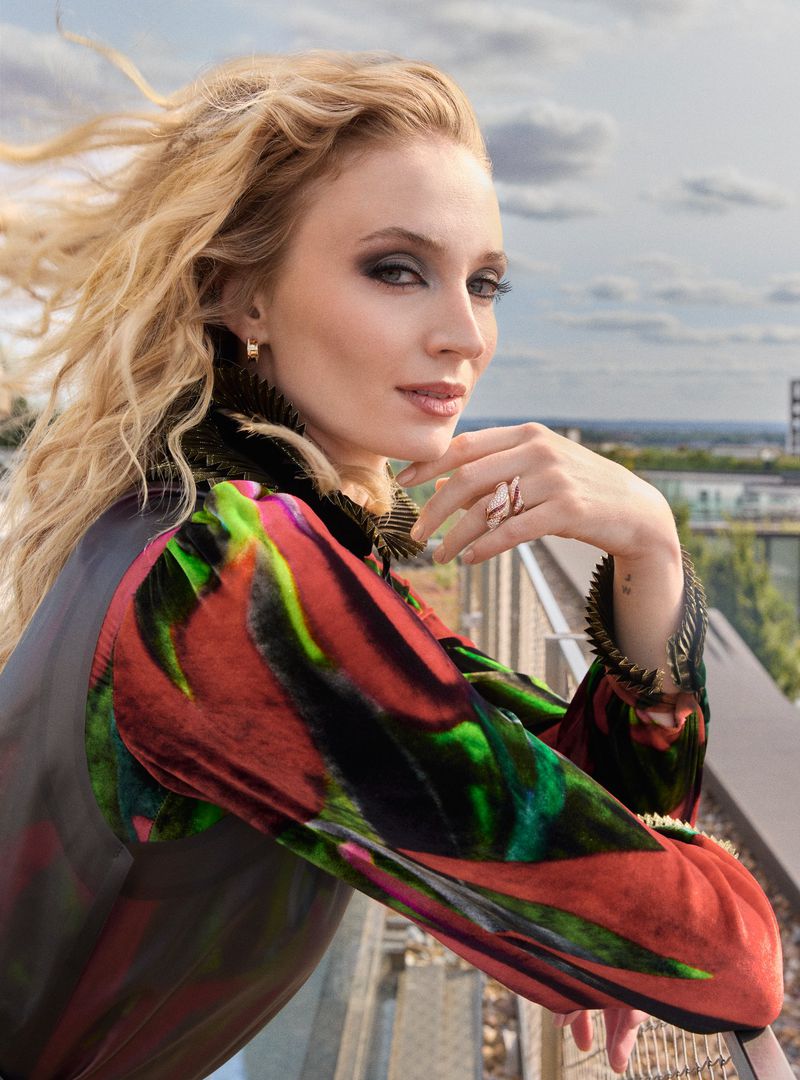 Portrait of a blonde woman in a colorful patterned top standing on a rooftop with a blurred cityscape and cloudy sky in the background.