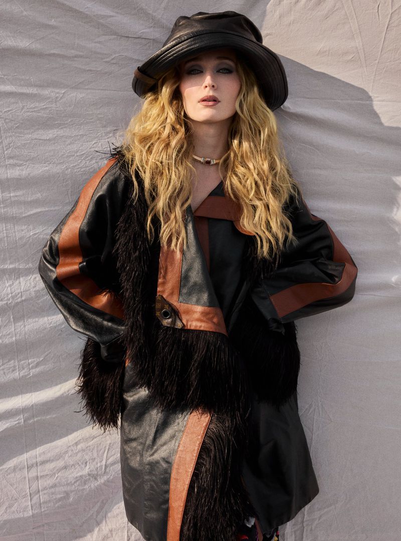 A woman in a black leather and faux fur coat and hat poses against a white backdrop, illuminated by bright, natural light.