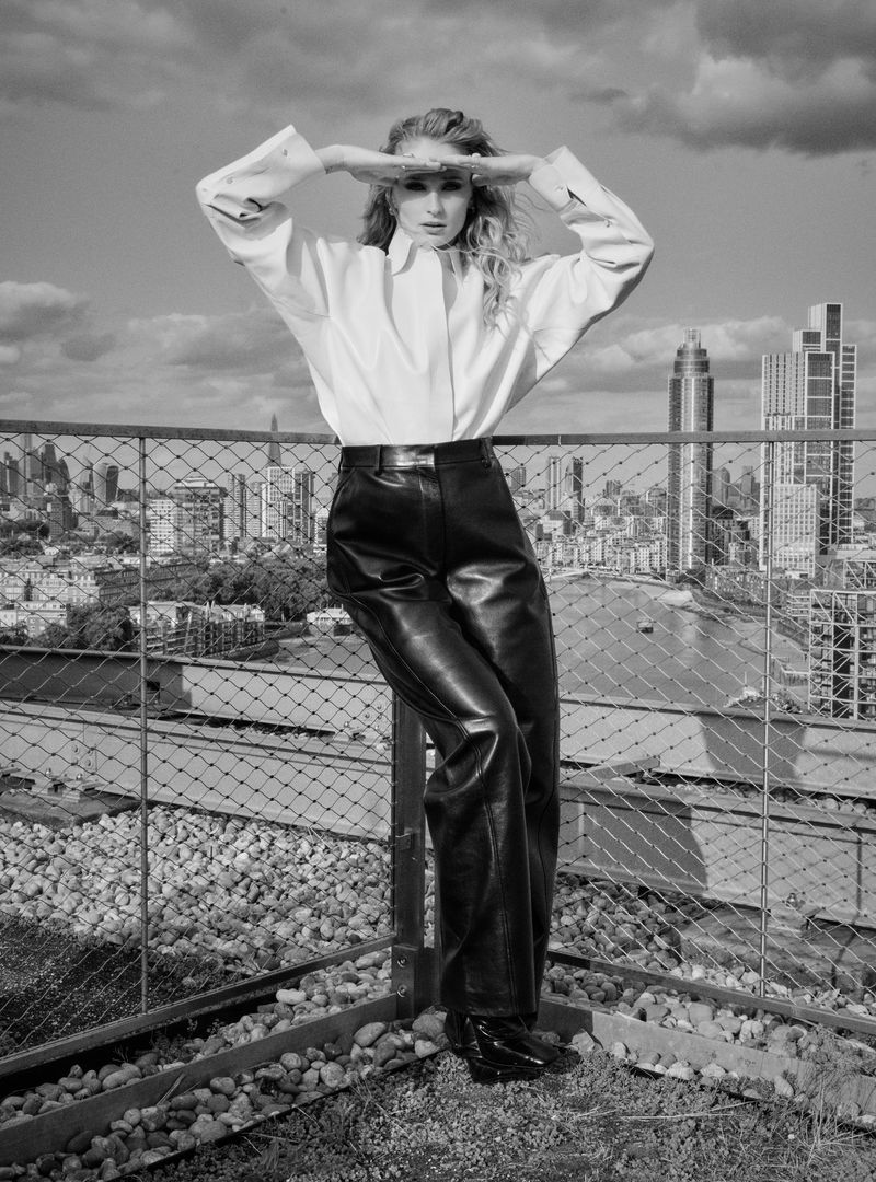 Black and white portrait of a woman in stylish clothing on a rooftop with a city skyline in the background on a clear day.