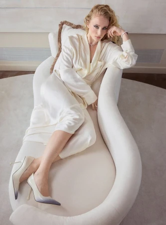 Adult female reclining on a white chaise lounge wearing a white robe and high heel shoes in an indoor setting.