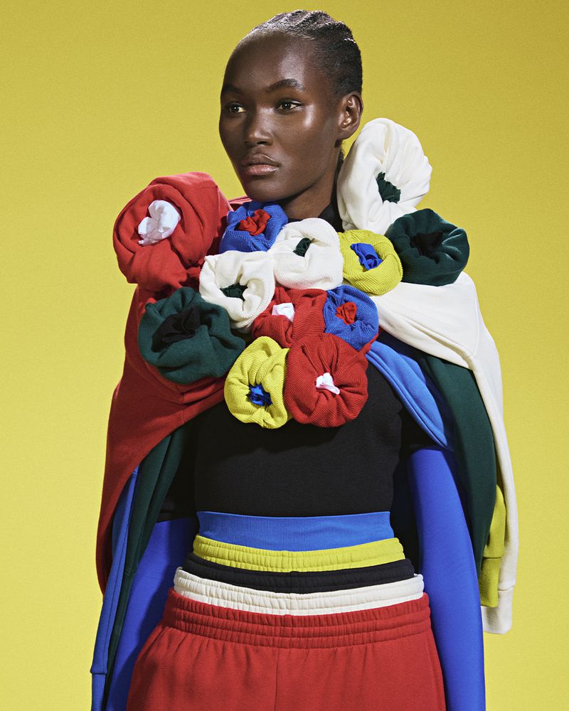 Dark-skinned woman in a black top and red pants posing in front of a yellow background, wearing a colorful garment of layered clothing.