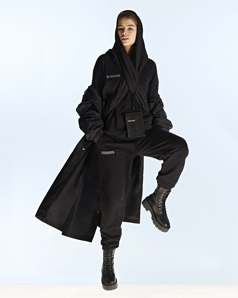 Woman in black hooded outfit and boots poses in a full shot on a light background, accessorized with a crossbody bag.