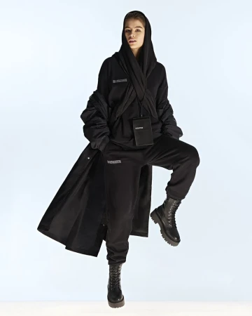 Woman in black hooded outfit and boots poses in a full shot on a light background, accessorized with a crossbody bag.