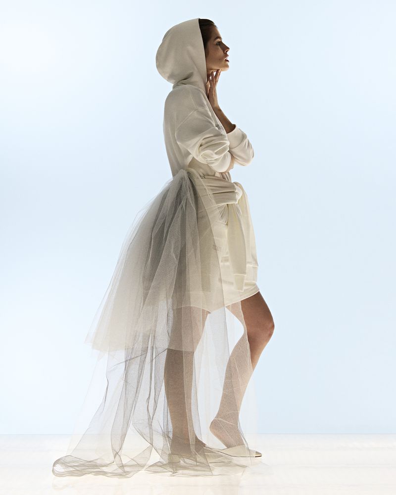 Woman in a white hooded dress with a tulle skirt, standing in profile against a soft blue background in a fashion style photograph.