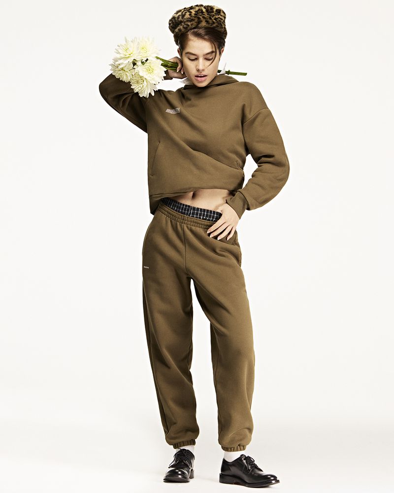 Full shot of a young adult woman in brown sweatsuit, leopard hat, and black shoes, holding white flowers against a white background.