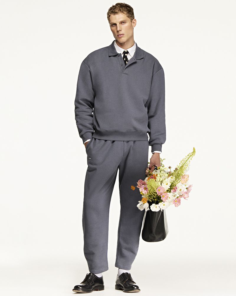 Male fashion model in gray sweatsuit holding flowers against a white backdrop, wearing black leather dress shoes and a tie.