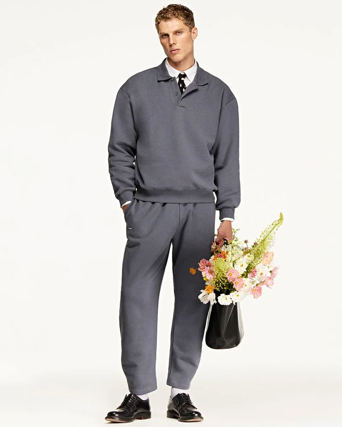 Man wearing a grey polo sweatshirt and matching trousers holding a bag of flowers against a white background for Pangaia