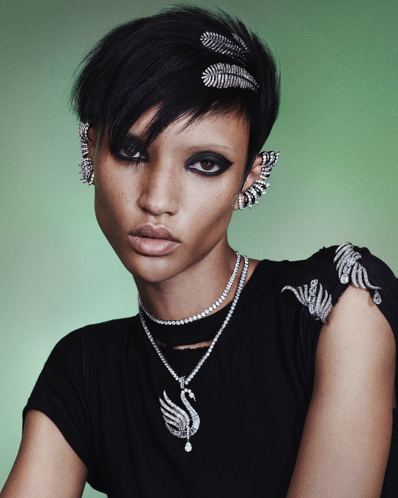 Close-up portrait of a young woman with short black hair and striking makeup, wearing elaborate jewelry against a green background.