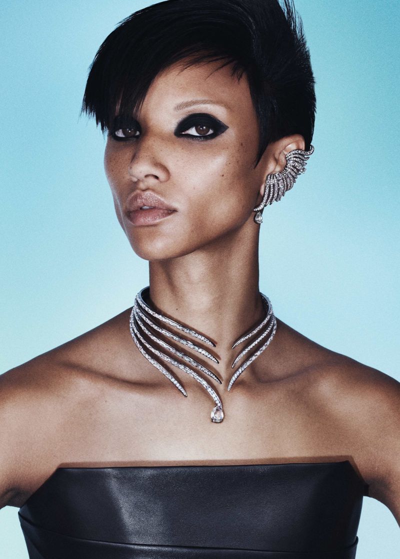 Close-up portrait of a woman with dark eye makeup wearing an ornate diamond necklace and matching ear cuff against a blue backdrop.