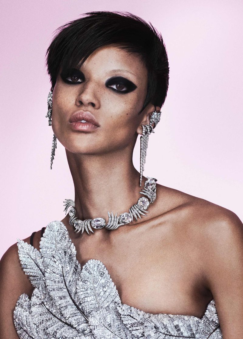 Close-up studio shot of a person with short black hair, dramatic eye makeup, silver statement jewelry, and a matching silver dress.