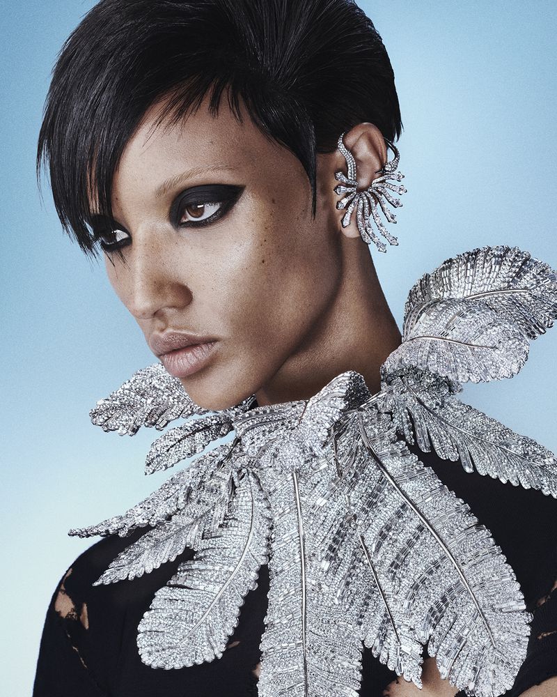 Close-up of a dark-skinned woman with short black hair, wearing a feather-like silver collar and matching ear cuff against a light blue background.