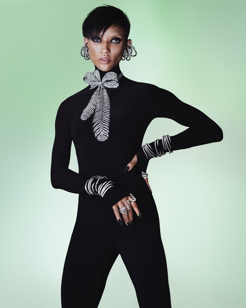 Portrait of an adult woman with black hair, wearing a black jumpsuit and silver jewellery, posing against a soft green gradient background.