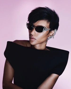 Model Georgia Palmer wearing crystal-embellished sunglasses and a wing ear cuff in a black structured top against a pink studio backdrop.