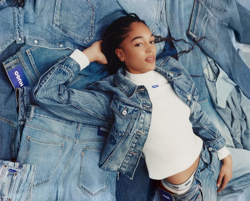 An adult woman with braided hair is lying down on a surface of light-blue jeans, wearing a light-blue denim jacket and a white shirt.
