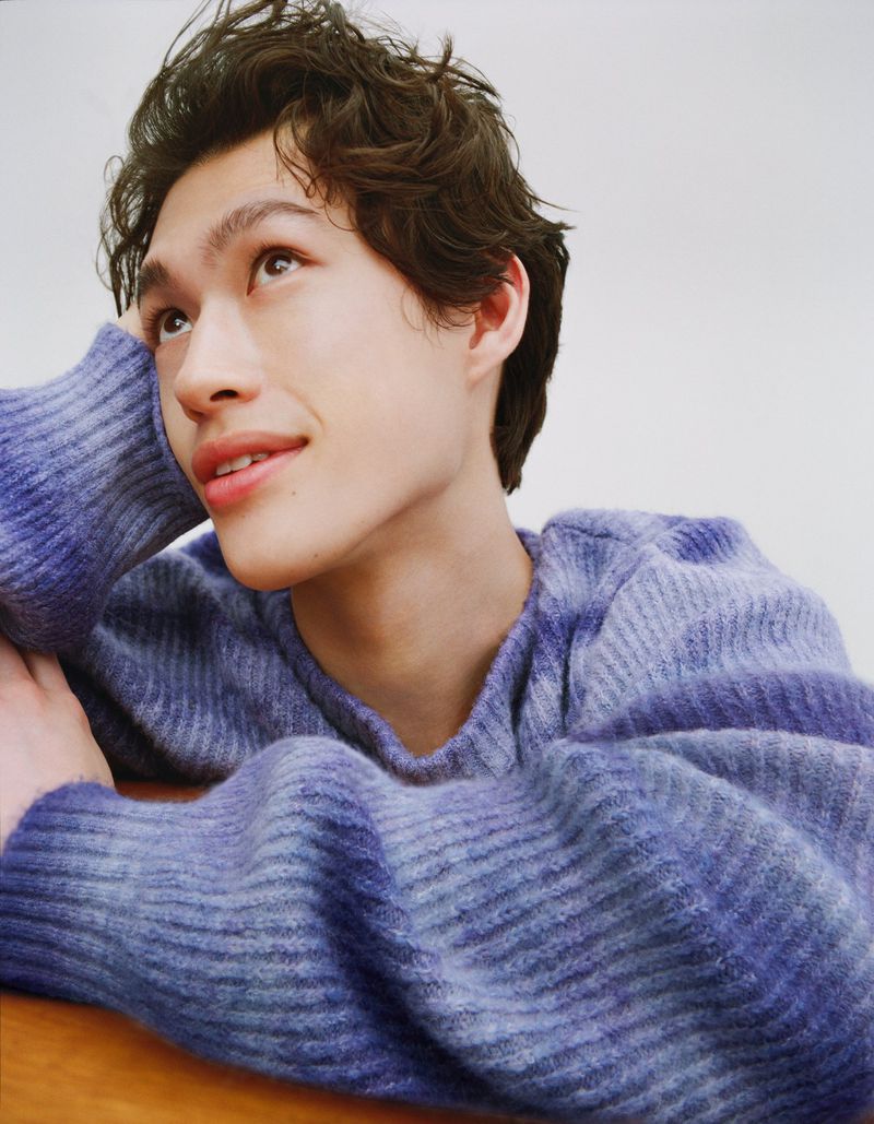 Portrait of a young adult with curly hair, wearing a blue and white sweater, looking up and smiling slightly, indoors against a white background.