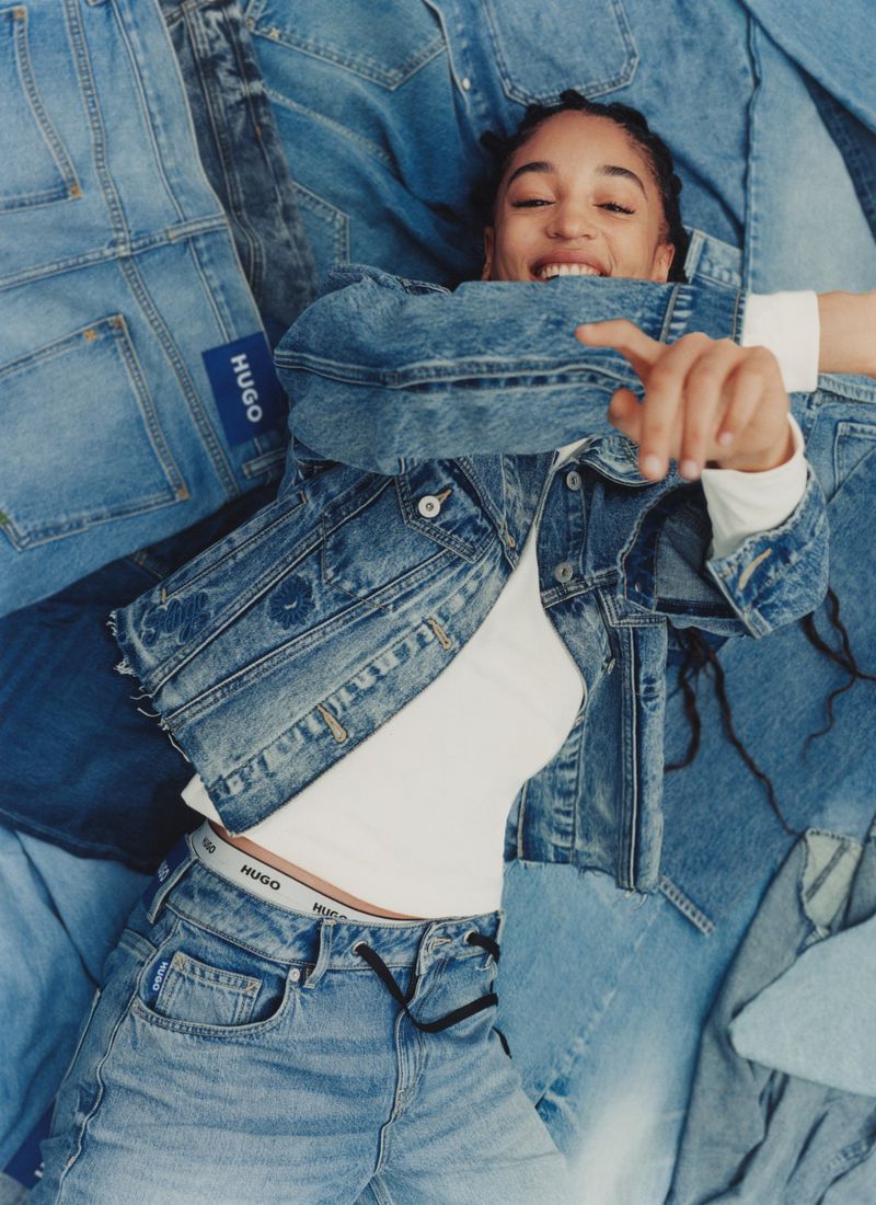 A fashionable young woman wearing denim clothes, including a jacket and jeans, smiles as she lies on a denim patchwork background.