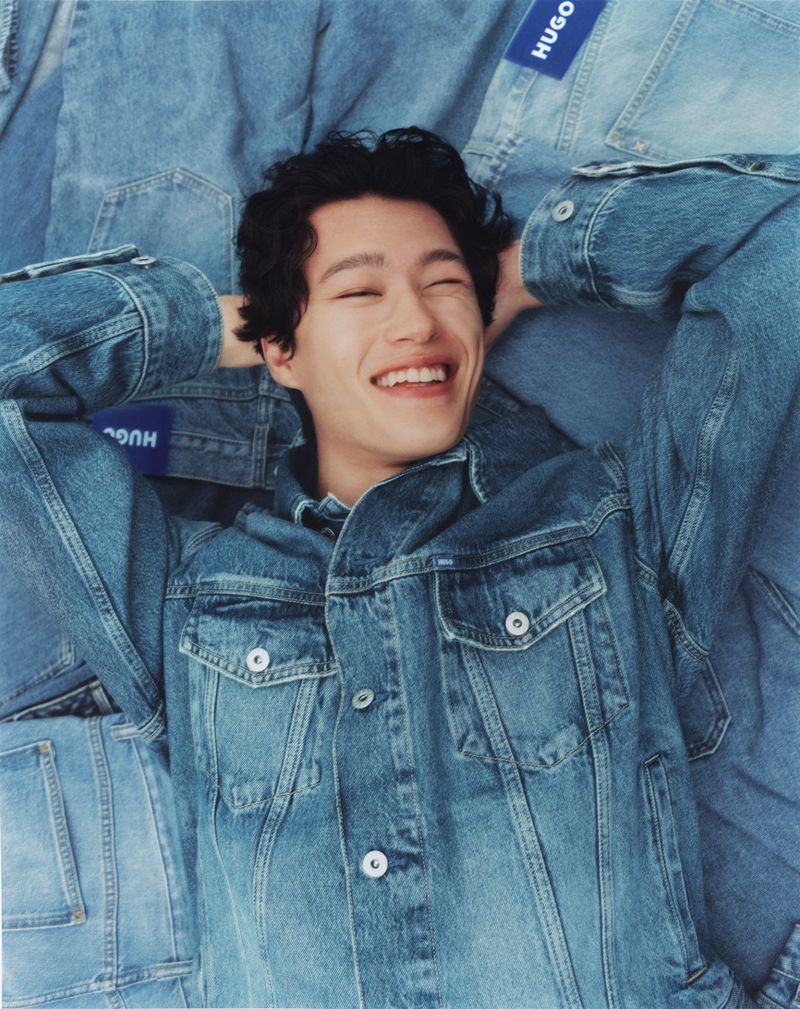 Smiling man with dark curly hair reclines on a pile of denim jackets, wearing a matching denim jacket. Soft light and a youthful vibe.