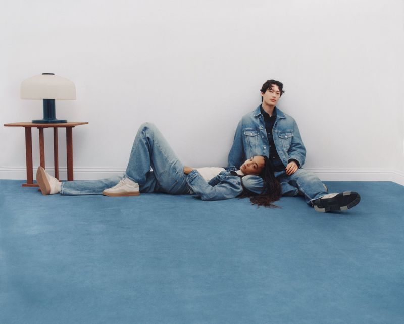 A young woman and man, casually dressed in denim, lying on a blue carpet in front of a white wall, with a modern table and lamp.