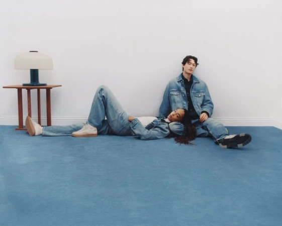 A young woman and man, casually dressed in denim, lying on a blue carpet in front of a white wall, with a modern table and lamp.