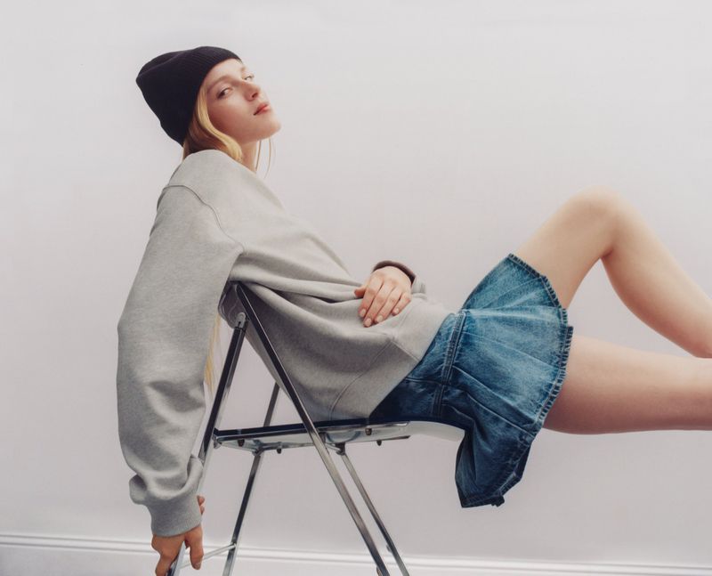 A young woman in a black beanie and grey sweatshirt sits on a folding chair in a studio wearing a denim skirt, captured with studio lighting.