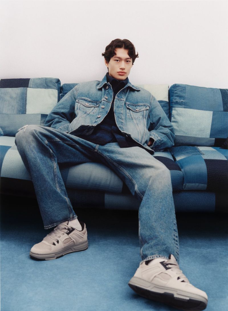 Man in denim jacket and jeans sits casually on a patchwork sofa, wearing sneakers, in a fashion forward casual pose indoors.