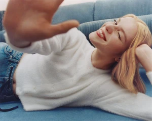 Woman in white fuzzy cardigan and denim jeans reclining on a blue sofa while raising her hand toward the camera