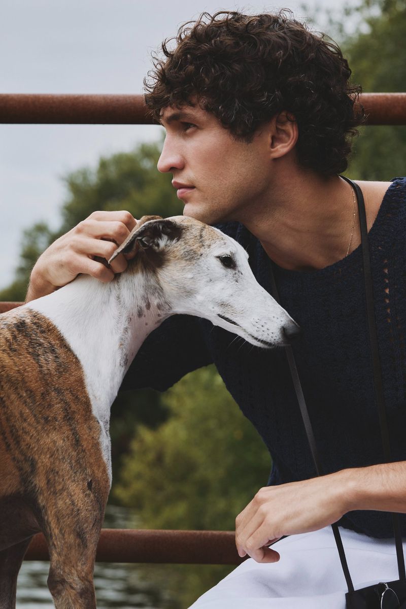 Young man poses with a white and brown brindle Greyhound dog outdoors; the man is petting the dog. Trees are visible in the background.