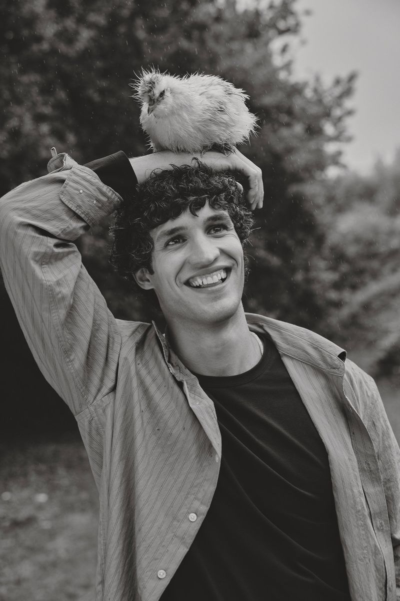 Smiling man with curly hair and button-down shirt in a nature setting, with a fluffy white chick perched on his head.