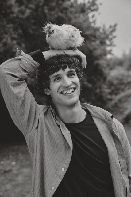 Smiling man with curly hair and button-down shirt in a nature setting, with a fluffy white chick perched on his head.