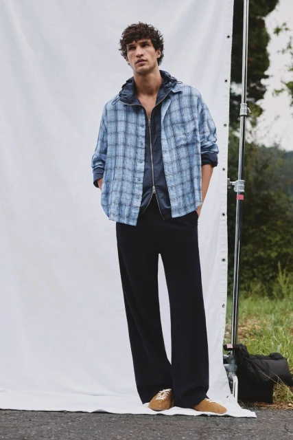 A fair-skinned adult male poses in a blue plaid shirt, dark jacket, black pants, and tan shoes against a white backdrop outdoors.