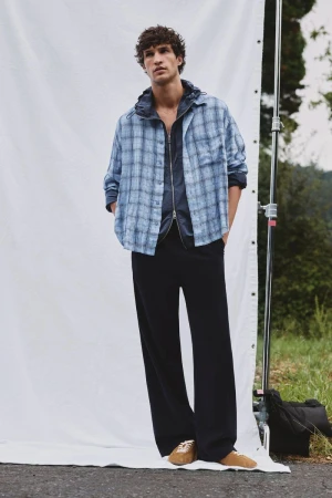 A fair-skinned adult male poses in a blue plaid shirt, dark jacket, black pants, and tan shoes against a white backdrop outdoors.