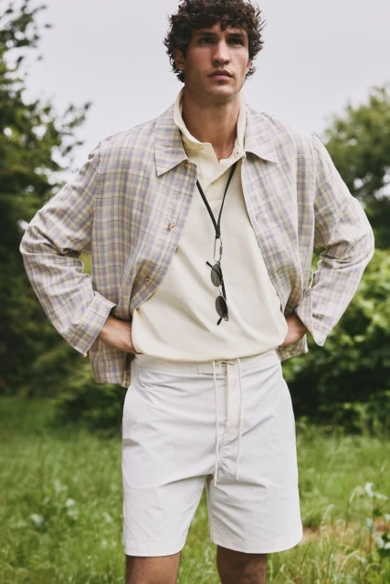 Adult male in a polo shirt and shorts stands outside wearing a plaid jacket with hands on hips in a medium shot.