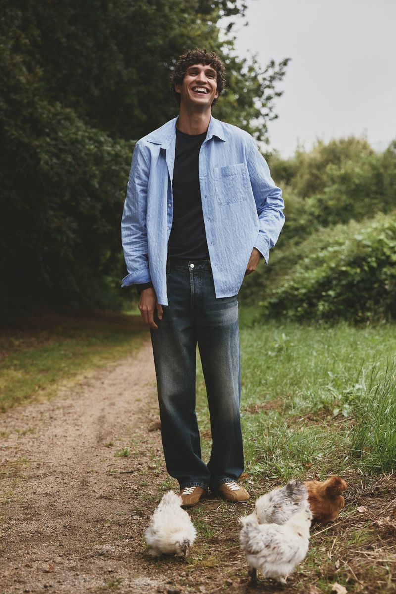 Man in unbuttoned light blue shirt and jeans standing on dirt path with chickens in grassy area with trees in the background.