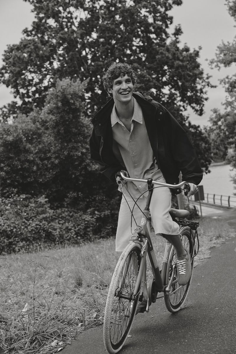 Monochrome portrait of a smiling man riding a bicycle on a path through a park, surrounded by trees and greenery in daylight.