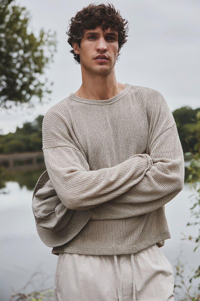 Man with curly brown hair in a beige long-sleeved shirt and pants stands outdoors with his arms crossed in front of a lake.