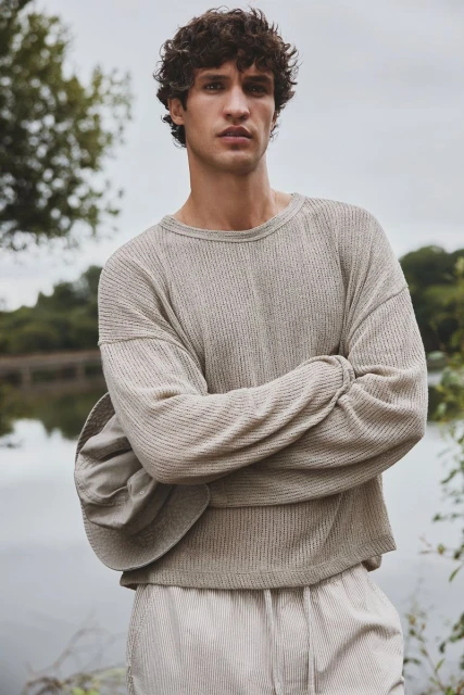 Man with curly brown hair in a beige long-sleeved shirt and pants stands outdoors with his arms crossed in front of a lake.