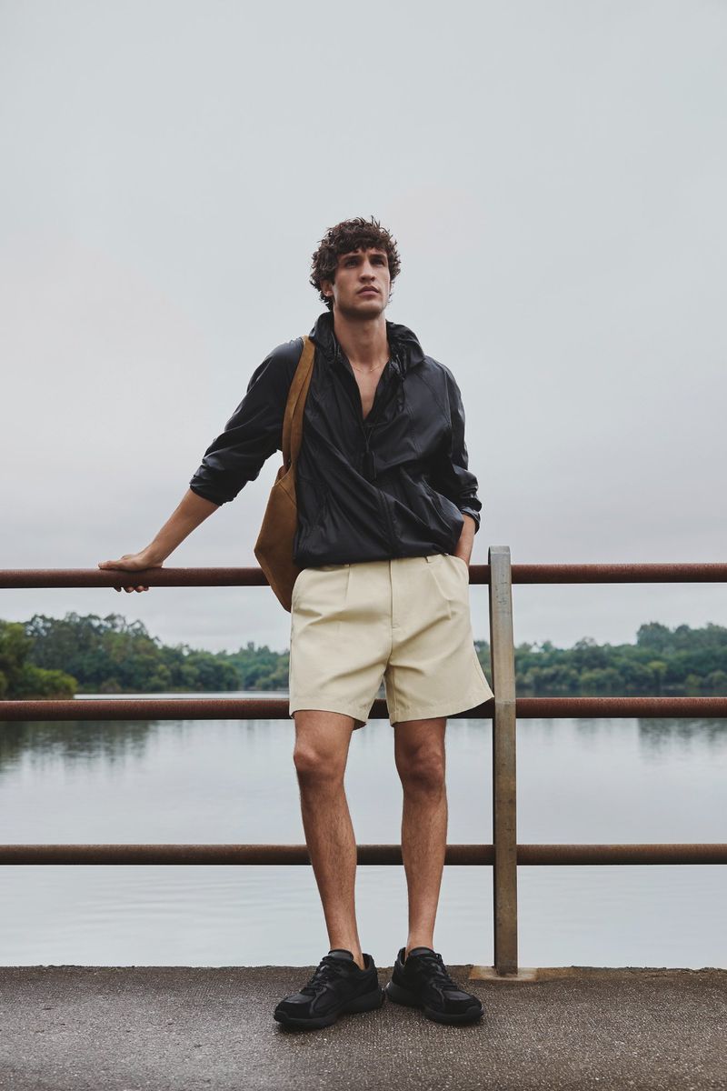 Francisco Henriques leaning on a metal railing near a lake, wearing a black lightweight jacket, cream shorts, and black sneakers.