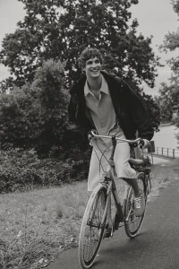 Model Francisco Henriques smiling while riding a bicycle down an outdoor paved path in a dark jacket and light shirt.
