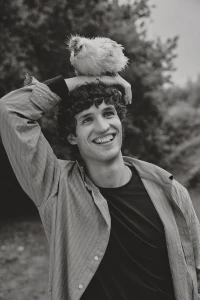 Smiling man holding a small fluffy bird on top of his head outdoors in a black and white editorial portrait.