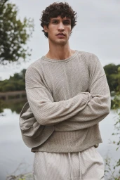 Male model with curly dark hair wearing a textured light beige sweater holding a hat outdoors by a river